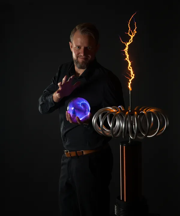 Thomas Sandner mit leuchtender Plasmakugel neben einer Teslaspule mit orangem Blitz