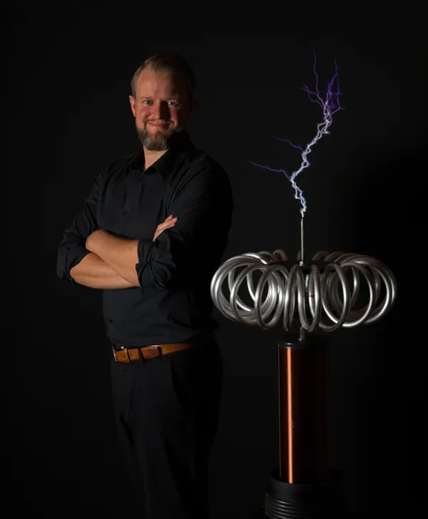 Thomas Sandner mit verschränkten Armen neben einer Teslaspule mit violettem Blitz